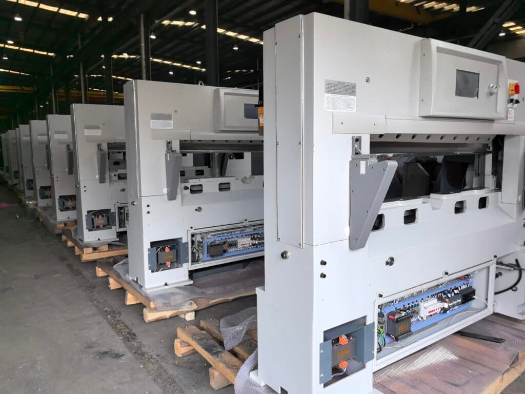 Row of new hydraulic paper cutting machines in warehouse with electrical control panels open for inspection and wiring verification.
