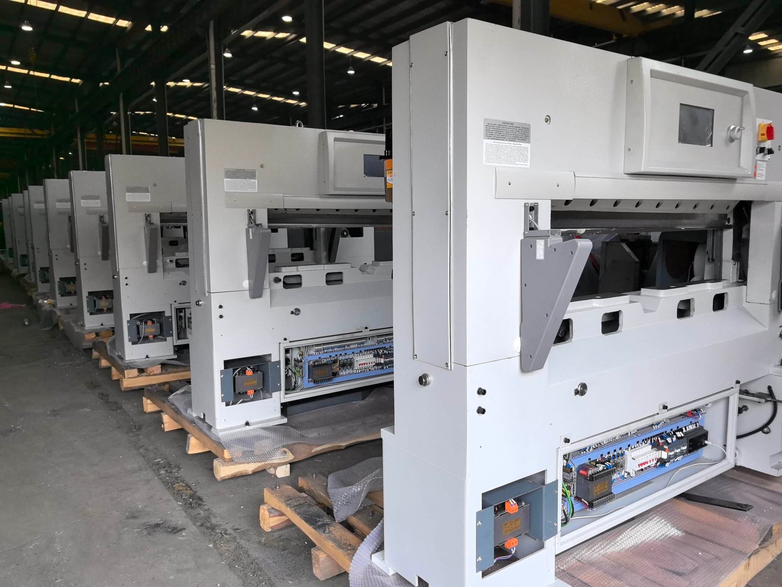 Row of new hydraulic paper cutting machines in warehouse with electrical control panels open for inspection and wiring verification.