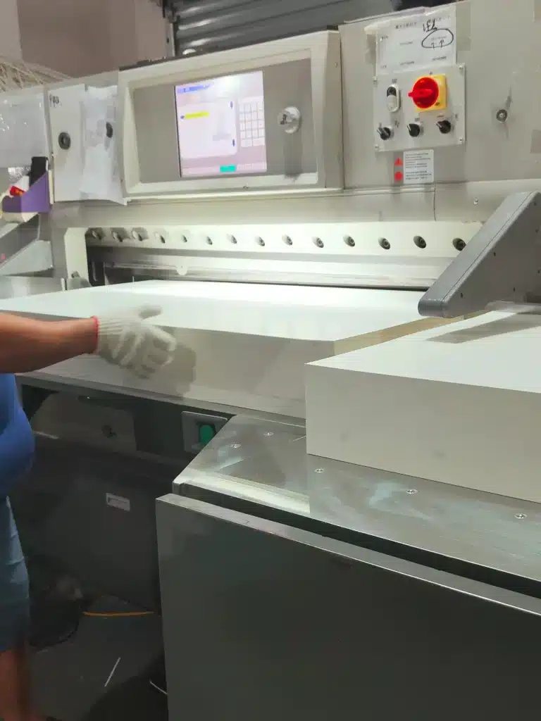 Worker handling thick paper stack on a hydraulic paper cutting machine emphasizing material stability during cut cycle