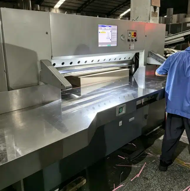 Heavy-duty guillotine cutter in operation with visible waste trimmings from thick packaging stock