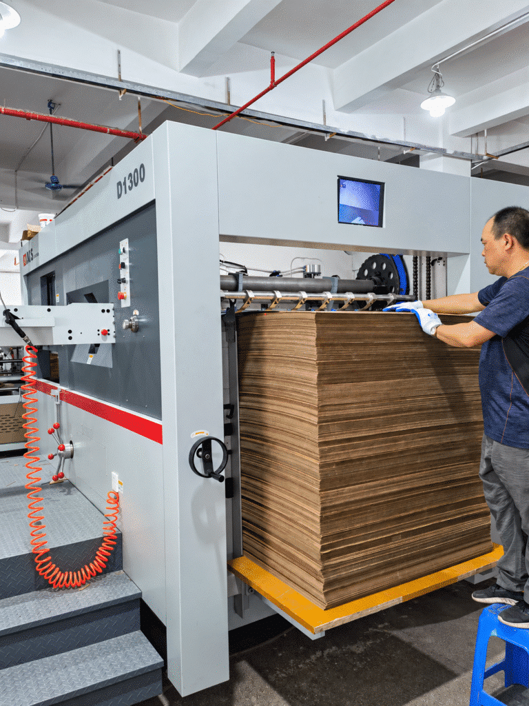Operator documenting job-cycle data on a clipboard beside a semi-automatic die cutter with finished blanks and production sheets visible