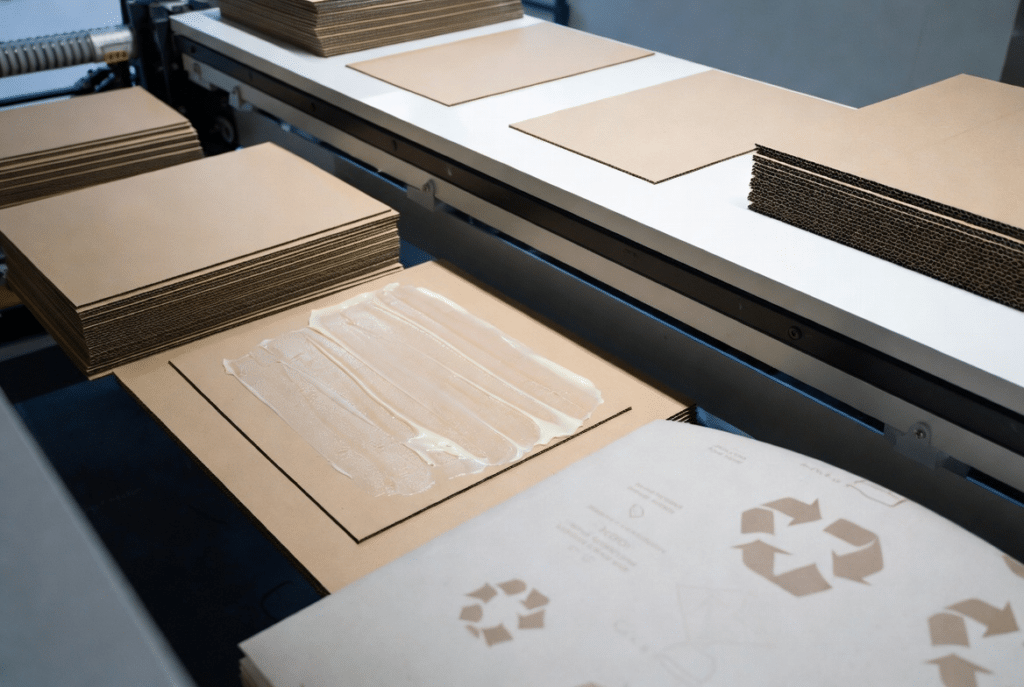 Industrial view of automatic laminating equipment applying precise adhesive to printed paperboard and corrugated sheets in a clean packaging factory, illustrating sustainable adhesive selection for EU and retailer recyclable packaging requirements