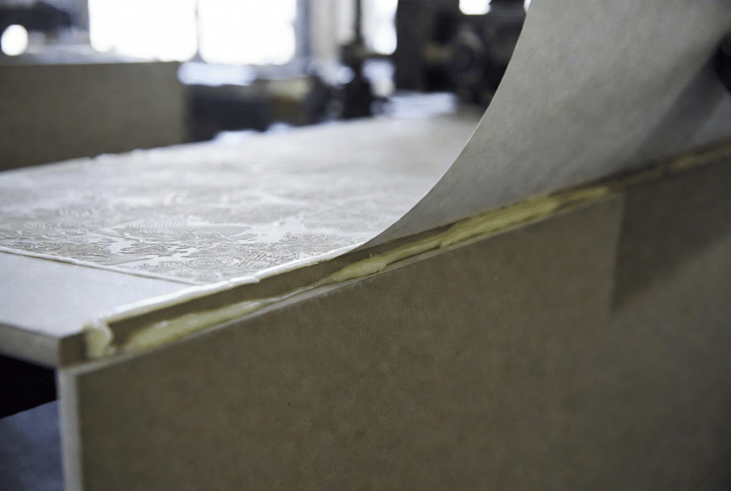 Decorative printed paper being positioned and bonded onto dense greyboard panel with visible glue layer during rigid box paper lamination process in packaging factory